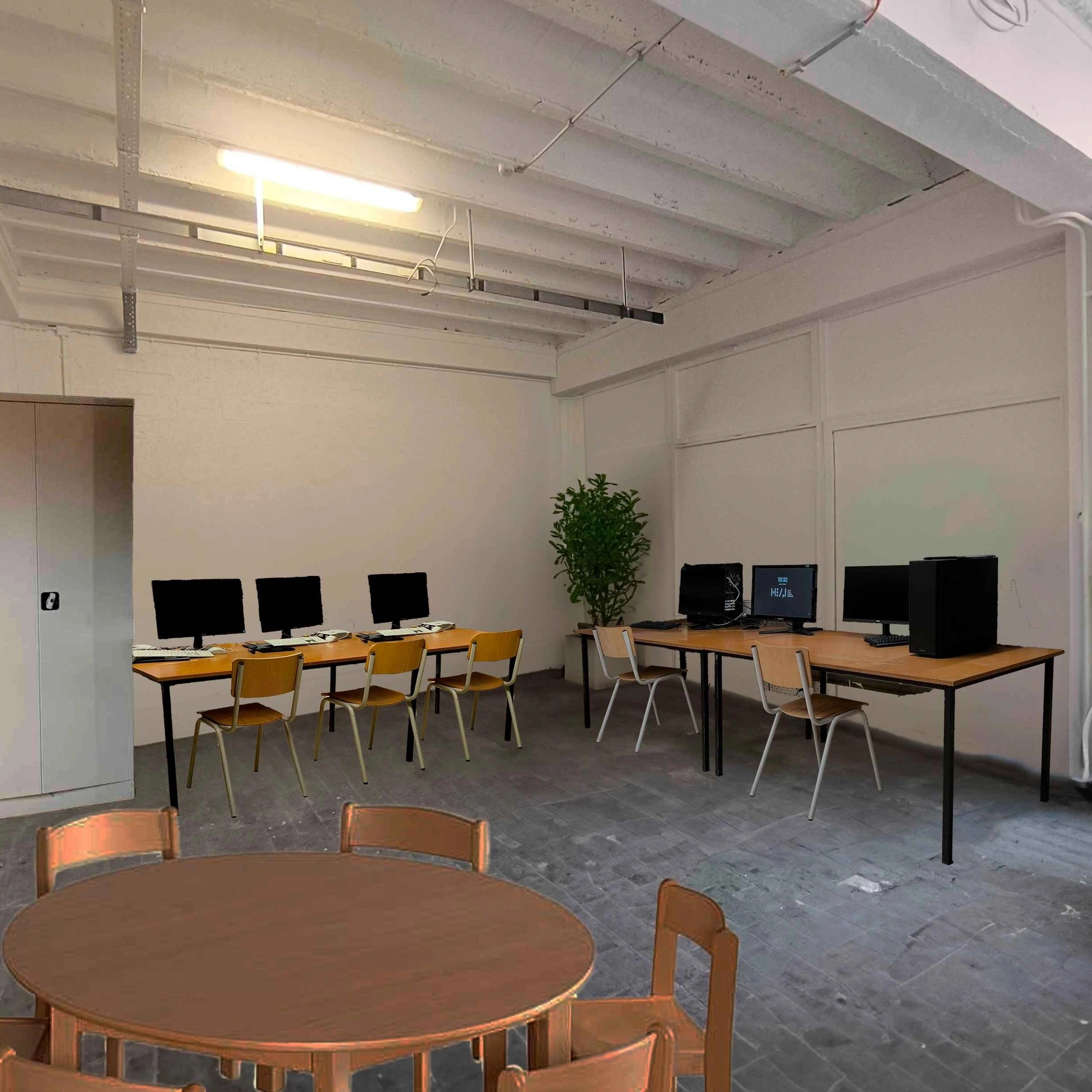 Image retouchée montrant les aménagements du mobilier dans le coin gauche opposé à la porte d'entrée. Le long du mur à gauche, on retrouve une table avec des ordinateurs et une armoire metallique. LE long du mur perpendiculaire on voit une autre table sur laquelle il y  aussi des ordinateurs. Dans l'angle des 2 murs, on voit une plante. Au premier plan, on voit une table ronde et des chaises (partiellement visible).